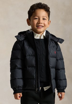 Ragazzo sorridente che indossa una giacca a piumino nera, un maglione nero lavorato a trecce e una camicia bianca con colletto su uno sfondo semplice.