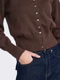 Bruine gebreide cardigan met een geribbelde zoom en manchetten, met knopen aan de voorkant. Draagt donkere blauwe spijkerbroek, met één hand in de zak.