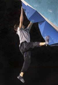 Osoba drži veliki plavi držač za penjanje na boulder zidu, odjevena je u sivu majicu i crne hlače za penjanje, s crnim penjačkim cipelama.