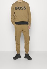 Man som bär beige cargojoggers och matchande sweatshirt med "BOSS" tryckt i stora svarta bokstäver, kombinerat med svarta sneakers och svart tröja under.