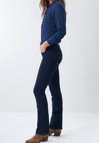 Chemise en denim à boutons avec deux poches poitrine, manches longues et col classique, associée à un jean évasé bleu foncé et des bottines brunes.