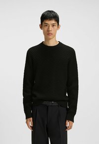 Junger Mann, der steht, trägt einen strukturierten schwarzen Strickpullover und schwarze Hose mit einem schwarzen Gürtel, vor einem schlichten hellen Hintergrund.