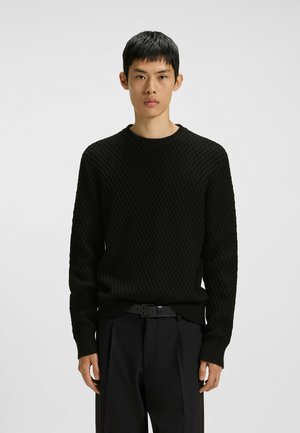 Un jeune homme debout, portant un pull en tricot noir texturé et un pantalon noir avec une ceinture noire, sur un fond clair simple.
