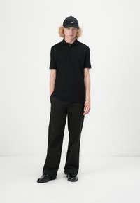 Svart polo-skjorte med teksturert mønster, kombinert med løse svarte bukser. Modellen har på seg en svart caps med en liten logo og svarte sko.