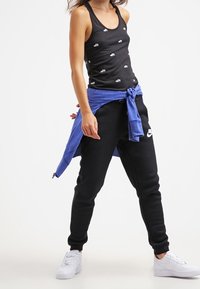Black tank top with small white logos, paired with black sweatpants and a blue hoodie tied at the waist, completed with white sneakers.