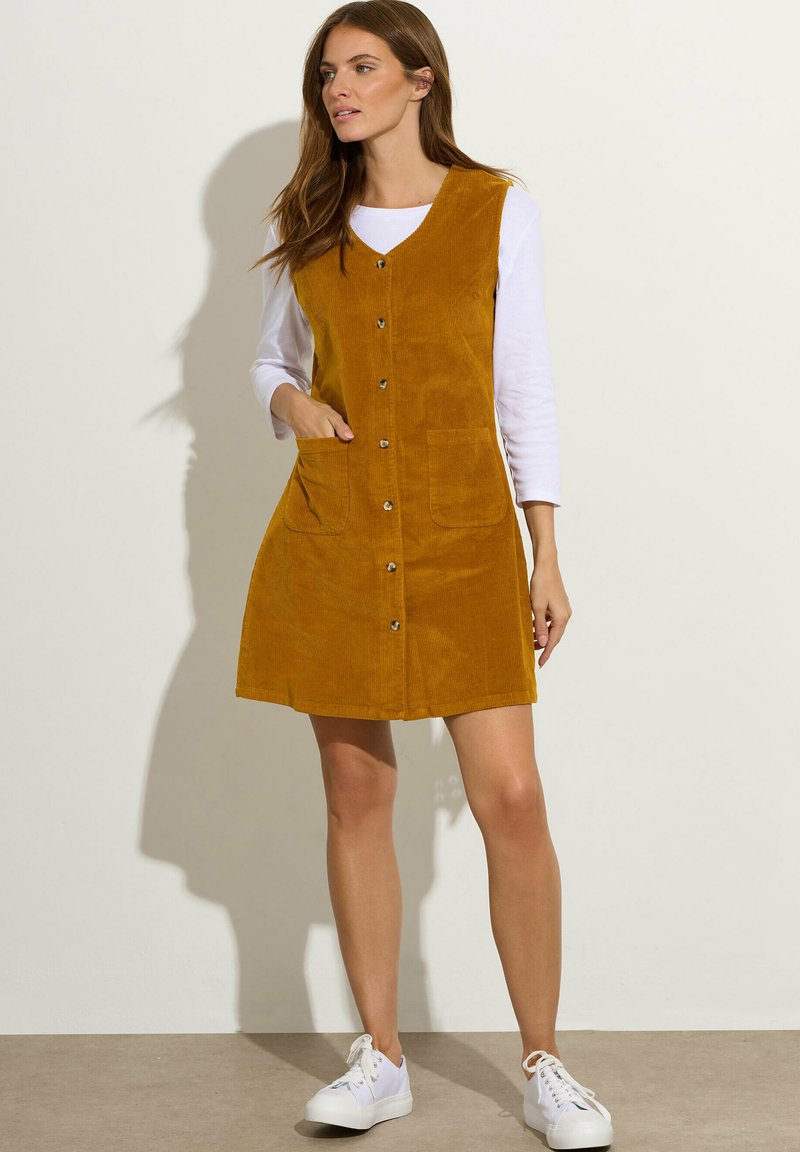 Vestido de pana mostaza con escote en V, botones en el frente y dos bolsillos de parche. Combinado con una camisa blanca de manga larga y zapatillas blancas.