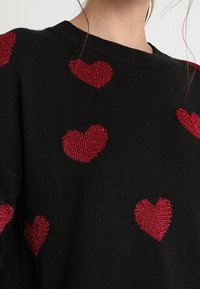 Musta neulepaita, jossa on sinne tänne sijoitettuja punaisia sydänkuvioita. Sydämet ovat teksturoituja ja metallinhohtoisia, mikä tuo niihin erottuvan visuaalisen yksityiskohdan.