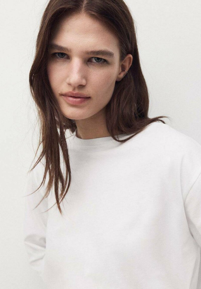 Biała koszulka z długim rękawem i okrągłym dekoltem. Modelka ma proste brązowe włosy i neutralny wyraz twarzy. Miękka, gładka faktura materiału.