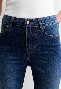 Jeans de mezclilla azul oscuro con un diseño ajustado, bolsillos frontales y un cierre de botón con un acento de cobre. Detalles sutiles de textura y costura visibles.