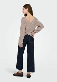 Pull tricoté beige clair avec un dos en V, associé à un jean large bleu foncé et des chaussures marron à lacets avec des semelles épaisses.
