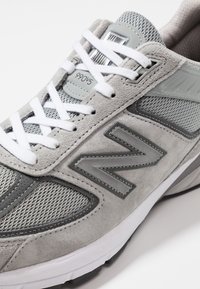 Baskets New Balance 990v5 grises et blanches avec des panneaux en daim et en mesh, des lacets blancs et un grand logo "N" sur le côté.