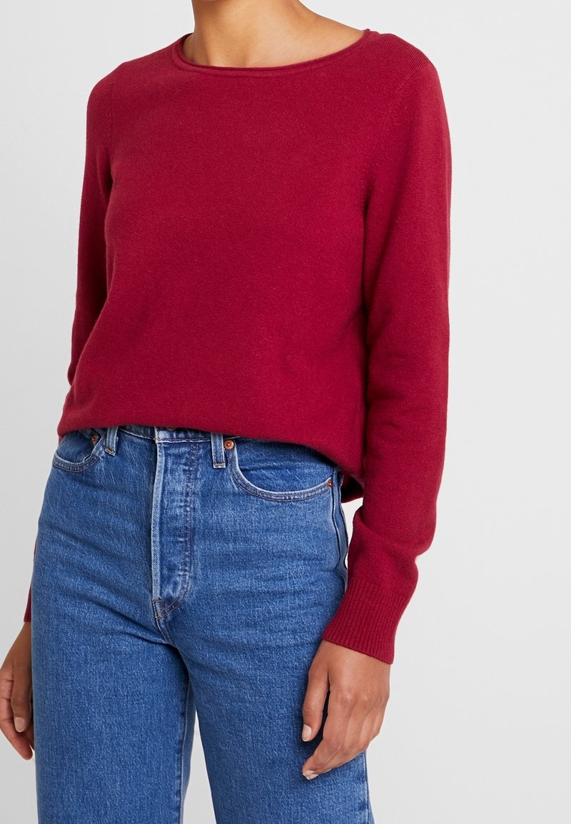 Roter Strickpullover mit rundem Ausschnitt und gerippten Bündchen, kombiniert mit hoch taillierter blauer Jeans. Glatte Textur und figurnaher Schnitt.