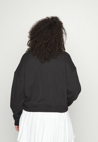 Melnas pārāk lielas izmēra džemperis, izgatavots no mīksta auduma, ar nokareniem pleciem un ribotām apakšmalām, redzams no aizmugures. Zem tā ir balta svārku.