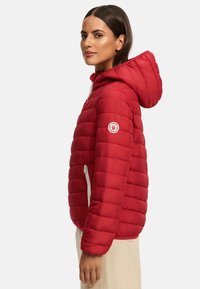 Red puffer jacket with a hood, featuring horizontal quilted design, a white zipper, and a circular logo patch on the upper arm.