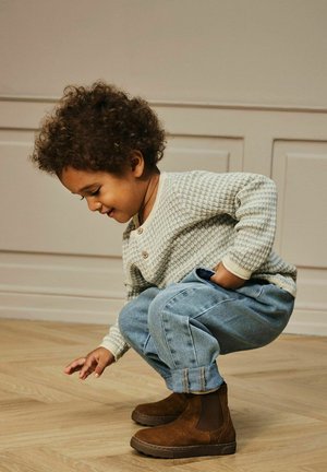Un bambino piccolo con capelli ricci accovacciato, che indossa un maglione leggero a fantasie, jeans arrotolati e stivali marroni su un pavimento in legno all'interno.