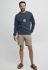 Marineblå hettegenser med et sirkulært logo og tekst, parret med beige shorts og brune sandaler, med en avslappet passform og teksturert materiale.