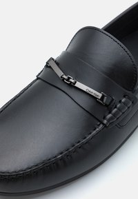 Sorte skinn lofer med en elegant design, en glatt tekstur, metallaksel med merkevare og detaljert søm langs kantene.