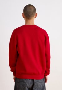 Person with short hair wearing a plain red sweatshirt and dark gray jeans, viewed from the back against a white background.