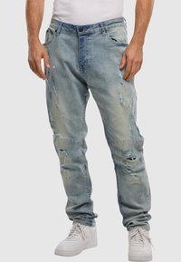 Jeans de mezclilla azul claro con detalles desgastados, con una cintura alta, bolsillos frontales y un diseño de pierna cónica. Llevados con zapatillas blancas.