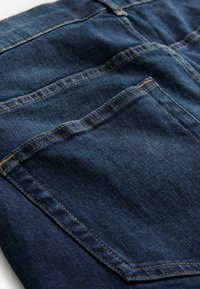 Țesătură de denim de culoare bleu închis, cu o textură netedă, evidențiind cusături vizibile și un buzunar din spate cu o cusătură diagonală.