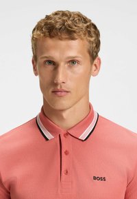 Rosa polo skjorta med ett texturerat tyg, klassisk krage med svart och vit accent, samt en liten svart logotyp på vänster bröst.