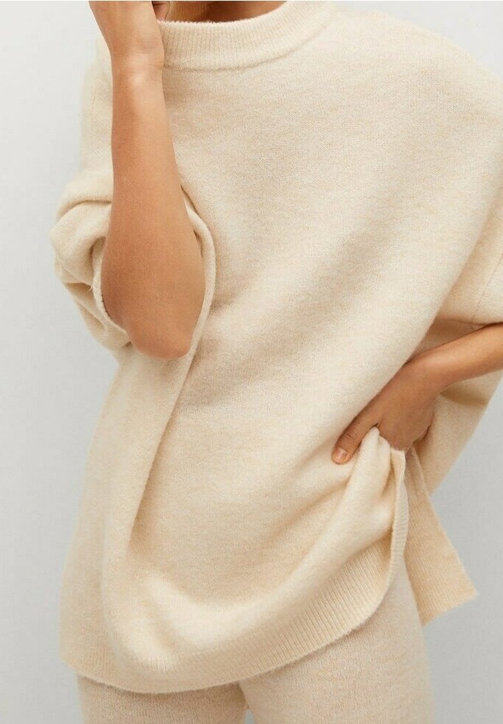 Personne portant un pull oversize beige doux et un pantalon assorti, une main tenant le côté du pull et l'autre près du visage.