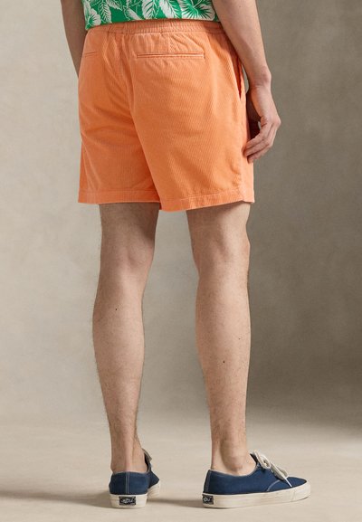 Πορτοκαλί σορτς από κοτλέ ύφασμα με υφή, ελαστική μέση και δύο πλαϊνές τσέπες, σε στυλ με πράσινο πουκάμισο με σχέδια και μπλε παπούτσια.