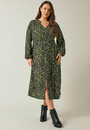 Langes schwarzes Kleid mit grünem und gelbem abstraktem Druck, V-Ausschnitt, geraffter Taille, langen Ärmeln und einem seitlichen Schlitz, kombiniert mit schwarzen Knöchelstiefeln.