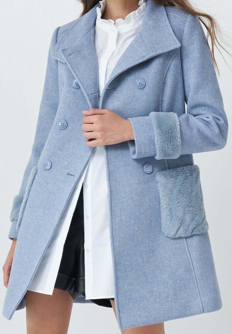 Donna che indossa un cappotto azzurro chiaro doppiopetto con polsini texturizzati e dettagli sulle tasche sopra una camicia bianca a bottoni e pantaloni corti neri.