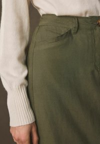 Pantalones de color verde oliva con una textura suave, con bolsillos laterales y cierre de botón, combinados con un suéter de manga acanalada blanca.