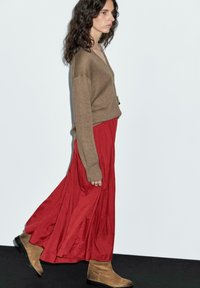 Pull en tricot marron, jupe rouge fluide et bottines beiges à la cheville. La tenue présente une coupe décontractée et un design superposé avec des textures douces.
