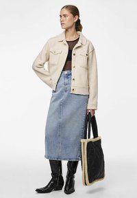 Veste en polaire beige avec deux poches poitrine, jupe en denim bleu, bottes noires à genoux, et un sac fourre-tout noir matelassé avec des bordures beiges.