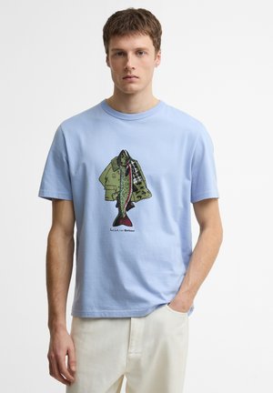 Jonge man die een lichtblauw T-shirt draagt met een afbeelding van een groene jas en een vis, gecombineerd met witte broek, staand tegen een eentonige achtergrond.