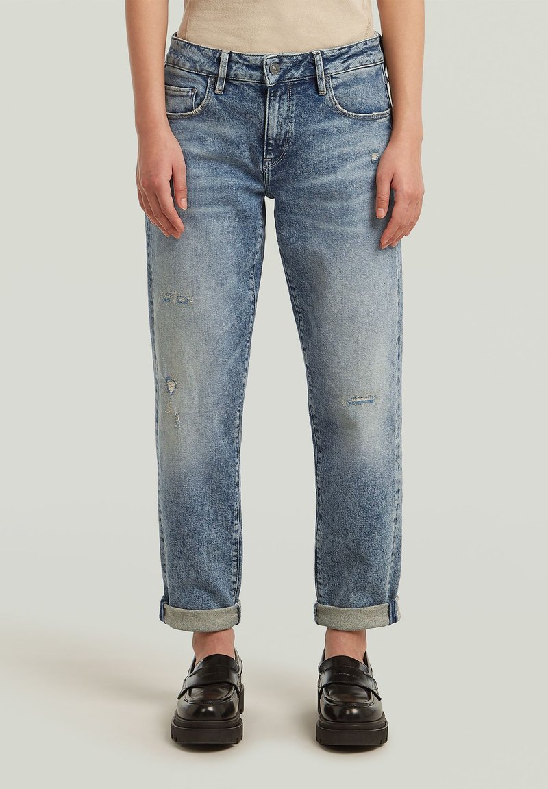 Jean en denim bleu clair avec une coupe décontractée, présentant des détails usés, des poignets roulés et un style classique à cinq poches. Des mocassins noirs portés en dessous.