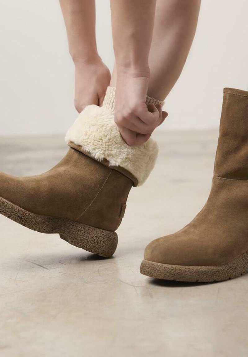 Botas de ante marrón hasta el tobillo con un dobladillo de piel de borrego, suela de goma texturizada y punta redonda. Persona ajustando el dobladillo con las manos.