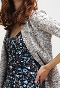 Grau strukturierter Cardigan über einem schwarzen Blumenkleid mit blauen Akzenten, der filigrane Muster und eine figurbetonte Silhouette zeigt.