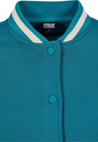 Jasnoniebieska kurtka z białym paskiem na ribonie i dwoma dopasowanymi zatrzaskami, metka mówi "Urban Classics XS, wyprodukowano w Chinach."