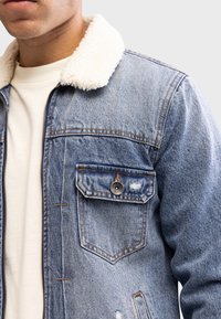 Kurtka dżinsowa z kołnierzem z baranka, w jasnoniebieskim kolorze, przednia kieszeń na guzik, przetarcia i widoczne przeszycia na ramionach i tułowie.