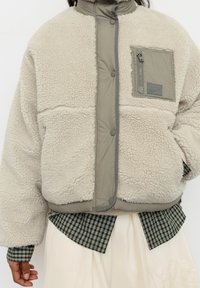 Fleecejakke i cremefarve med et polstret gråt panel og lynlåslomme. Har en høj krave og knaplukning samt en tekstureret overflade.