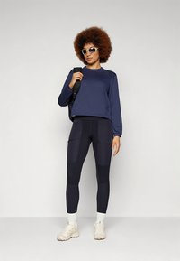 Sötétkék hosszú ujjú sportfelső, fekete zsebes legging nadrággal párosítva. Fehér zokni és bézs sportcipő egészíti ki az összhatást.