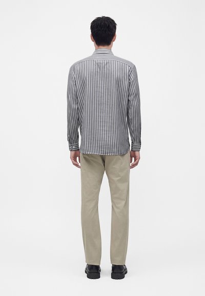 Homme debout de dos, portant une chemise à manches longues grise à rayures, un pantalon beige et des chaussures noires sur un fond blanc uni.
