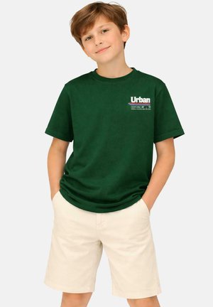 Jongen met lichtbruin haar die een donker groen T-shirt met het logo "Urban" en beige korte broek draagt, staand met handen in de zakken en een lichte glimlach.
