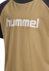 Tan katoenen t-shirt met zwarte raglanmouwen; heeft het witte logo "hummel" prominent op de voorkant. Soepele textuur.