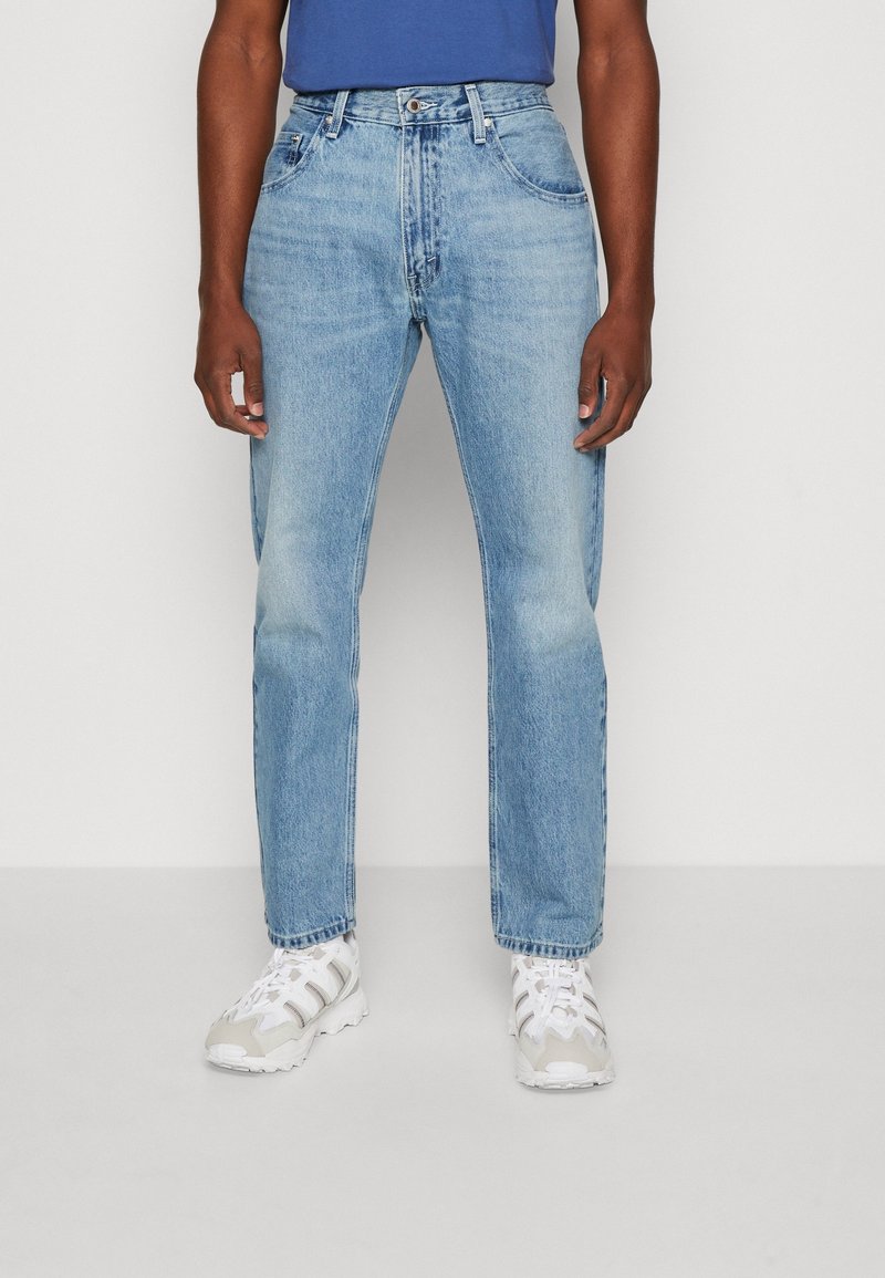 Jeans de mezclilla azul claro con un corte recto, cinco bolsillos y un cierre de botón, combinados con zapatillas blancas. Textura suave.