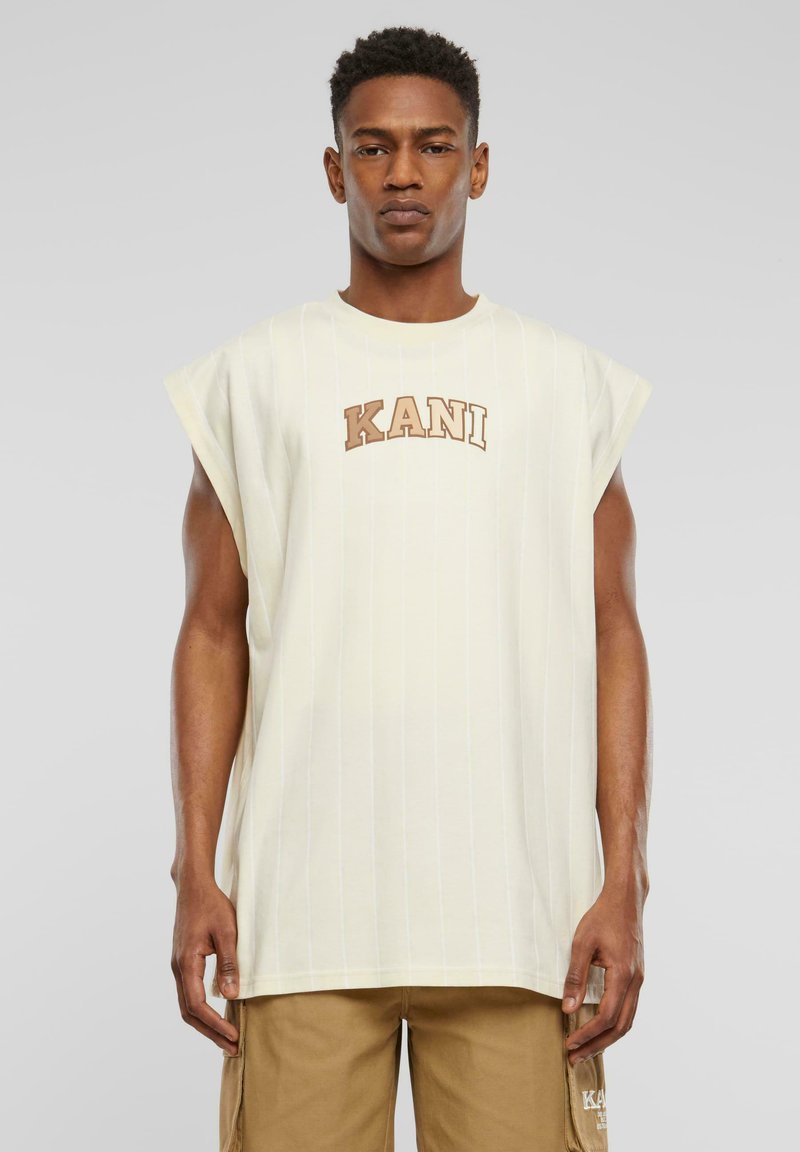 Camiseta sin mangas de gran tamaño de color claro, con rayas verticales y un logo "KANI" marrón en el pecho, fabricada en una tela suave.