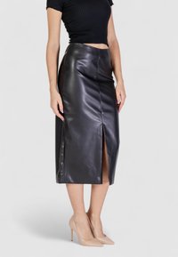 Falda midi de cuero negro con una abertura frontal, que presenta una textura suave y brillante y una silueta ajustada, combinada con tacones altos en tono nude.