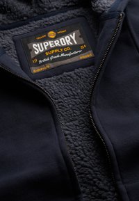 Marineblå zip-up hættetrøje med fleecefor, sort label med teksten "Superdry Supply Co." i gul og hvid, har metal lynlås.
