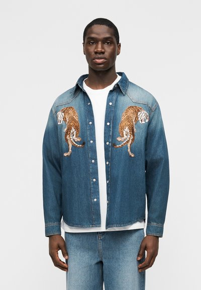 Νεαρός άνδρας που φορά ανοιχτό μπλε τζιν μπουφάν με δύο κεντημένα τίγρια στο στήθος, λευκό μπλουζάκι από κάτω και μπλε τζιν παντελόνι.