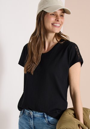 Lachende vrouw met een beige pet en een zwart T-shirt met kanten mouwen, houdt een bruin jasje vast, staand tegen een tweekleurige beige achtergrond.