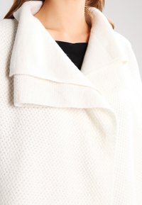 Vit strukturerad jacka med stor, vikbar krage, med ett texturerat tyg och en mjuk yta. Designen betonar värme och komfort.